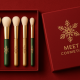 A red matte holiday gift box holds five makeup brushes with festive red, green, and gold handles, each stamped with a gold snowflake and topped with soft ivory bristles; beside the box sits the lid embossed in gold foil with a large snowflake and the words “MEET COSMETICS.”