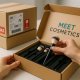 Hands removing a makeup brush from an open MEET COSMETICS branded cardboard box with black foam insert, while two stacked shipping boxes sit blurred in the background.